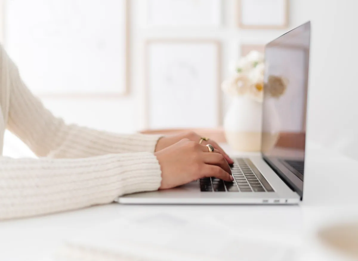 Close-up of a woman's hands typing on a laptop from the side and a flower vase in the background.