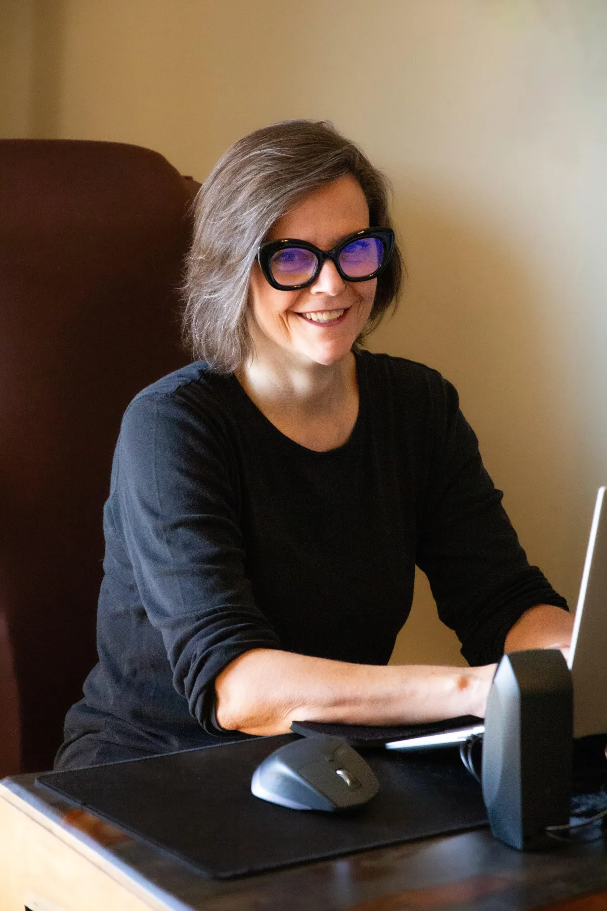 Women's book coach of memoir and nonfiction, Maurene Janiece, sitting at desk wearing black shirt and chunky glasses, smiling at the camera.