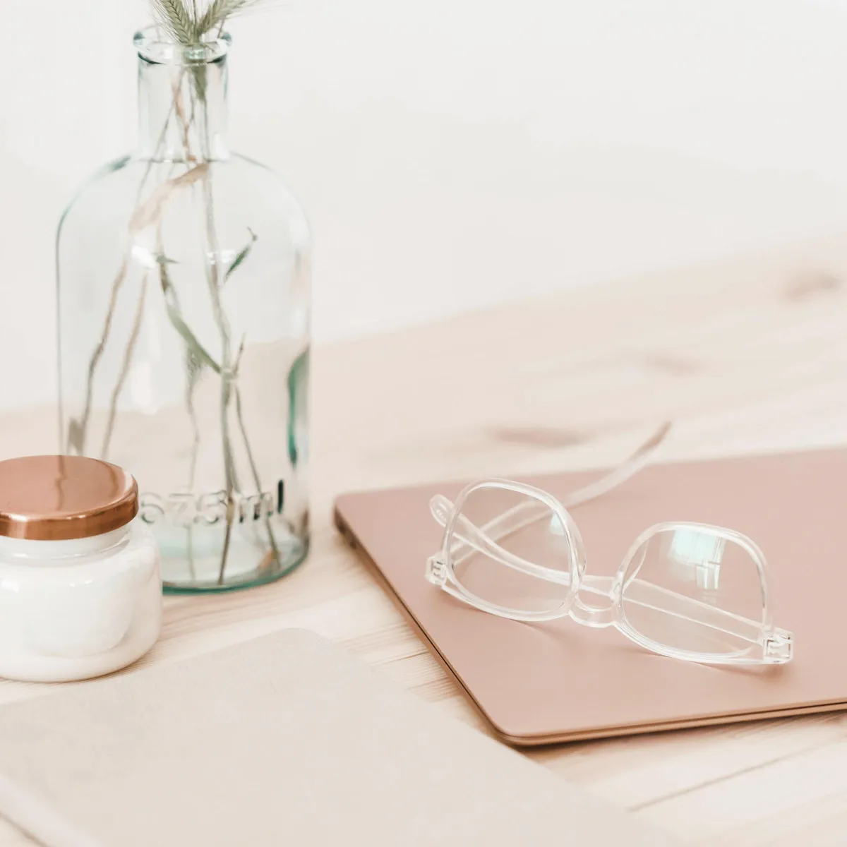 Close-up of reading glasses on top of a closed tan notebook and a white jar and clear vase.