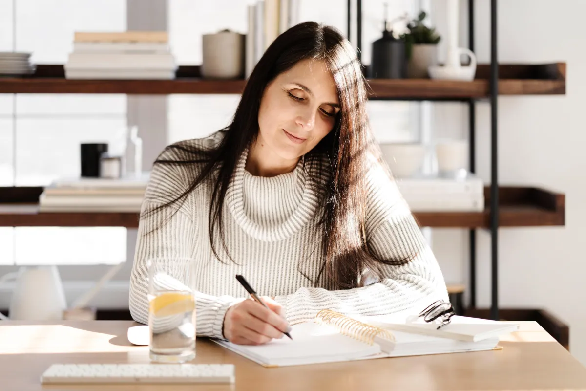 Midlife woman content while writing in an open notebook with sunshine coming in from the side.
