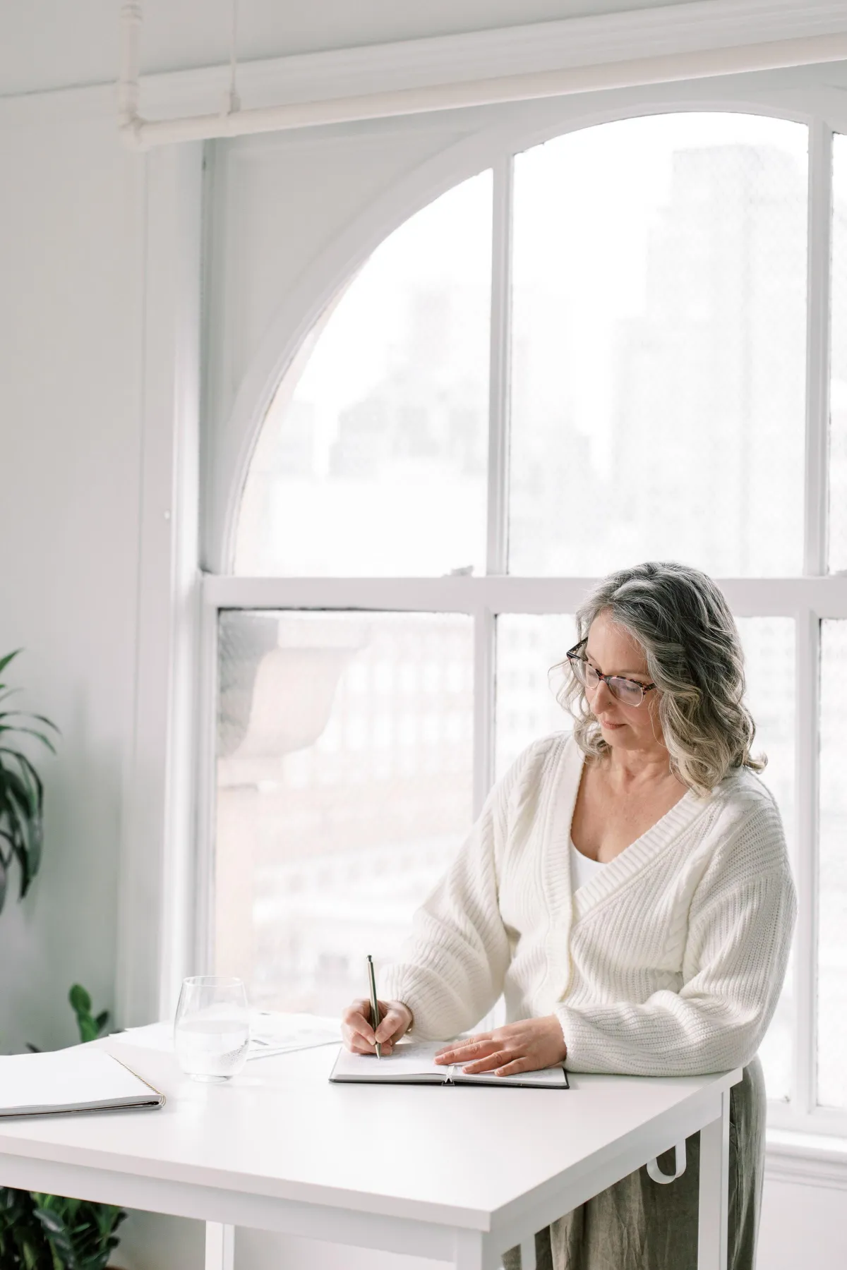 Mature woman standing at a table and writing in an open notebook looking content.