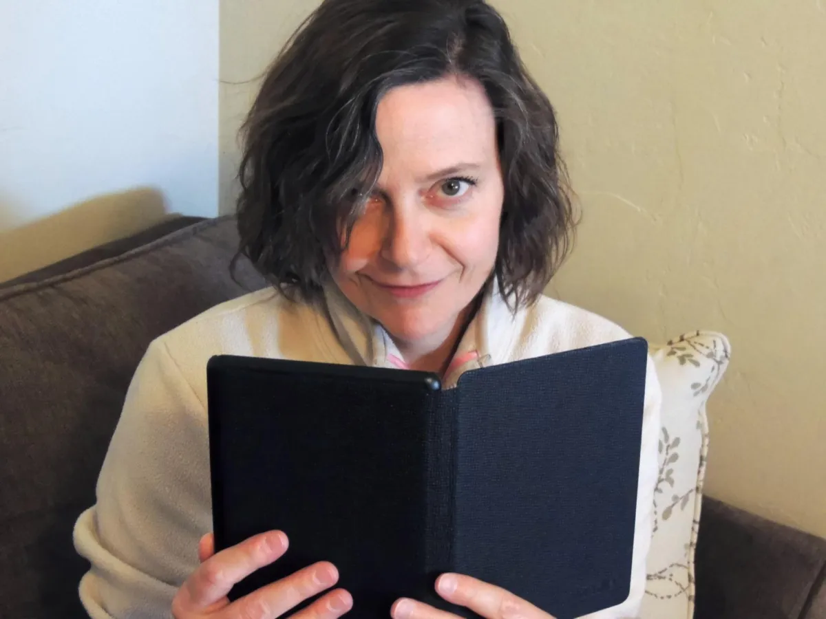 Book coach Maurene Janiece sitting on couch holding book and looking up at camera with a smile.