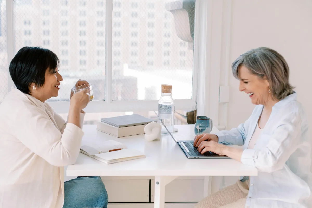 Two mature women sitting at a table laughing, one with an open notebook and the other typing on a laptop.