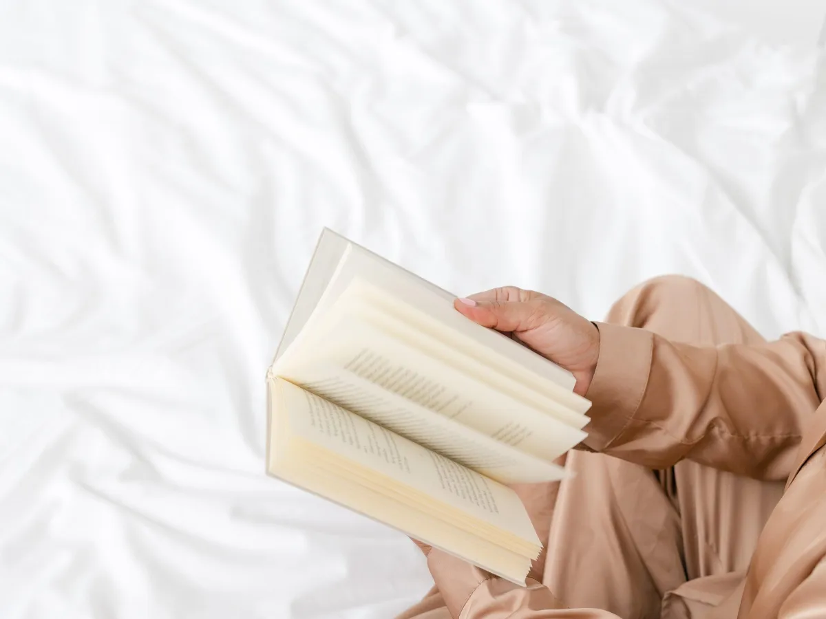A close-up of a woman sitting on a bed in tan pajamas flipping through a book with white fabric in the background.