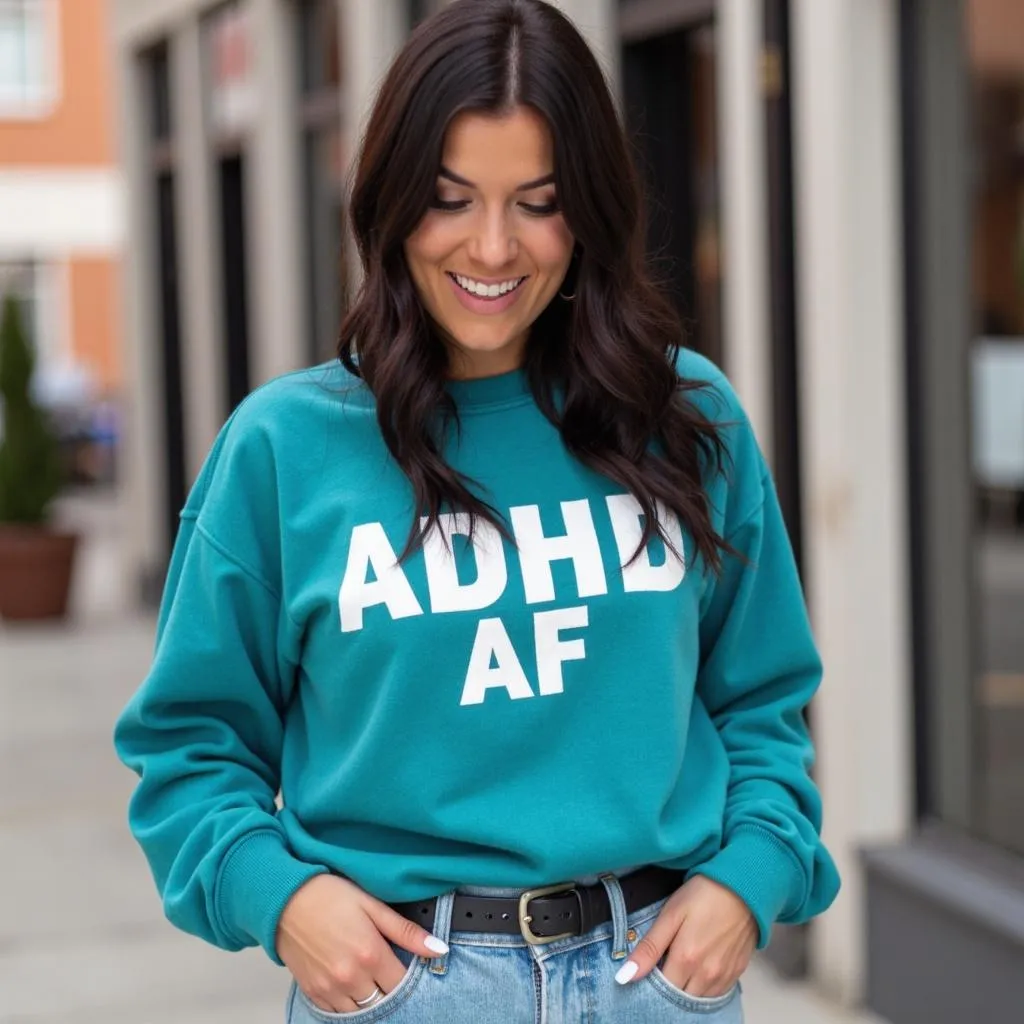 A woman with long, dark wavy hair smiles while looking down, wearing a teal sweatshirt with bold white text that reads "ADHD AF." She has her hands casually tucked into the front pockets of her high-waisted jeans, and the background shows an outdoor shopping or business area with blurred buildings and greenery.