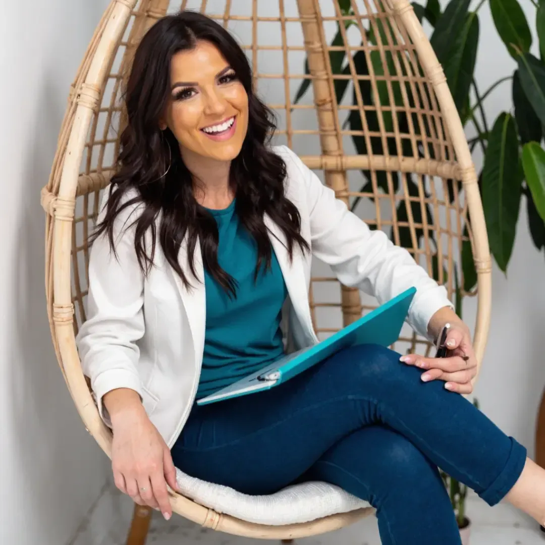 Alyece Smith, ADHD business mentor, smiling while seated in a wicker chair with a teal clipboard and pen in hand, wearing a white blazer.