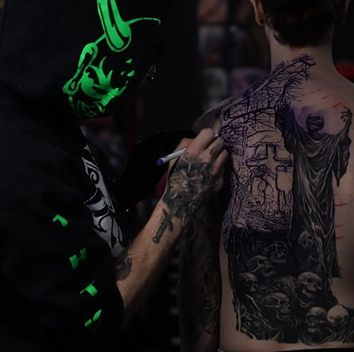 Black and grey realism and surrealism back tattoo in progress, featuring a dark religious scene with a robed figure and skulls, while a professional tattoo artist in Warren, Ohio maps the stencil and composition on a client’s upper back.