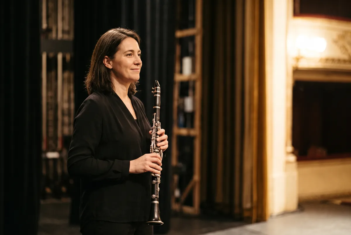 Clarinetista de pie entre bastidores, junto a las cortinas del escenario, sosteniendo su clarinete con una sonrisa serena; al fondo, los palcos iluminados de un teatro.