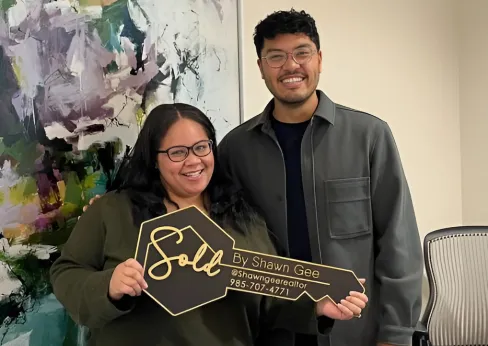 Shawn Gee with a happy client holding a 'Sold by Shawn Gee' key in front of a modern abstract art piece, celebrating a new home sale.