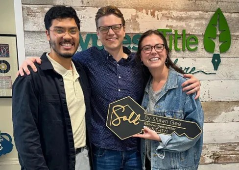 Shawn Gee celebrating with happy clients holding a 'Sold by Shawn Gee' key in front of the Synergy Title sign after purchasing their new home.