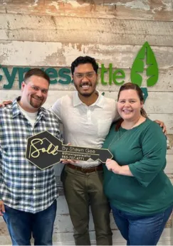 Shawn Gee smiling with a group of clients holding a 'Sold by Shawn Gee' key in front of the Synergy Title sign after closing a real estate transaction.