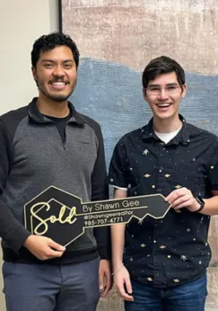 Shawn Gee posing with clients holding a 'Sold by Shawn Gee' key in front of a rustic wooden backdrop, marking a successful home sale.