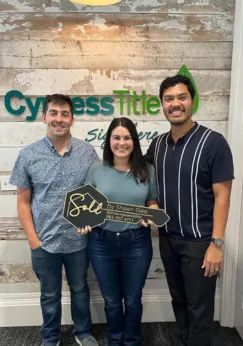 Shawn Gee with a couple holding a 'Sold by Shawn Gee' key in front of the Synergy Title sign, marking a completed real estate transaction.