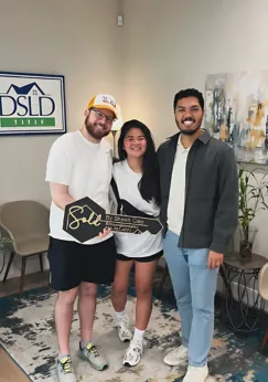 Shawn Gee posing with smiling clients holding a 'Sold by Shawn Gee' key in a bright office setting, celebrating their successful home purchase.