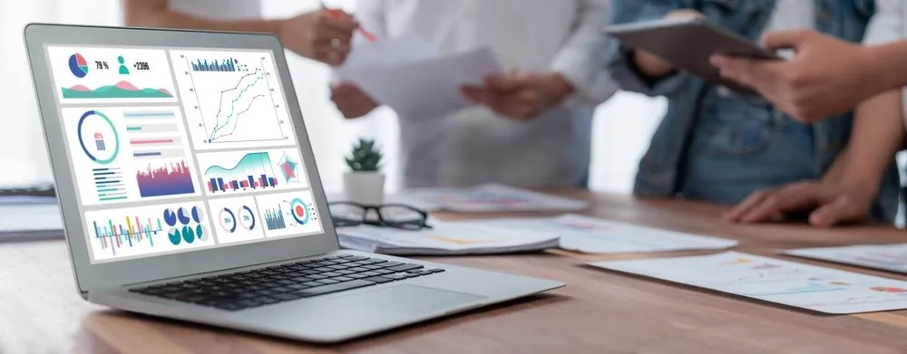 ChatGPT said:  Alt text: Open laptop displaying colorful data charts and graphs on a desk with people reviewing documents in the background.