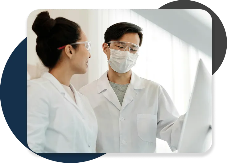 Two lab professionals in white coats reviewing data on a screen.