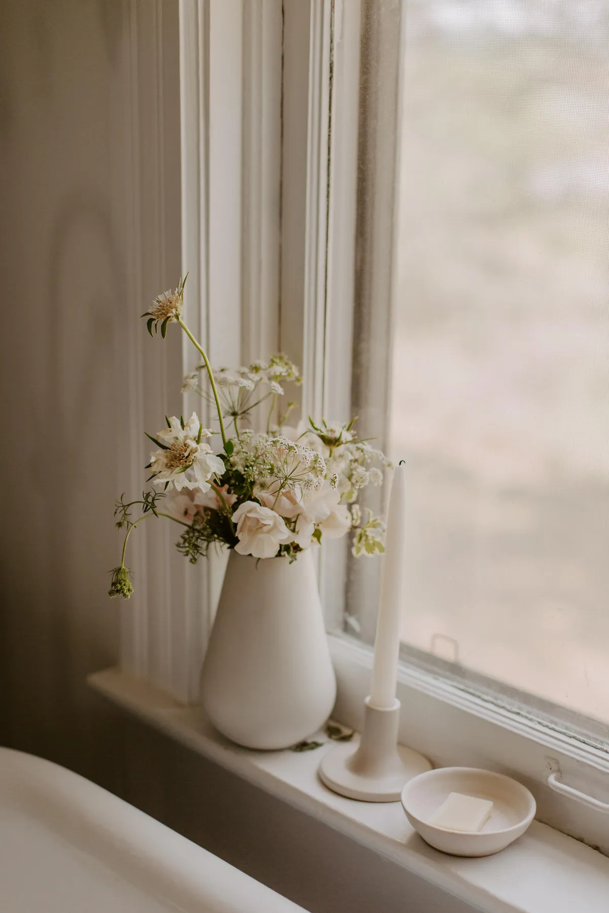 Soft floral arrangement and candle on a windowsill in a serene, light-filled home — calm, natural textures, and intentional sanctuary design
