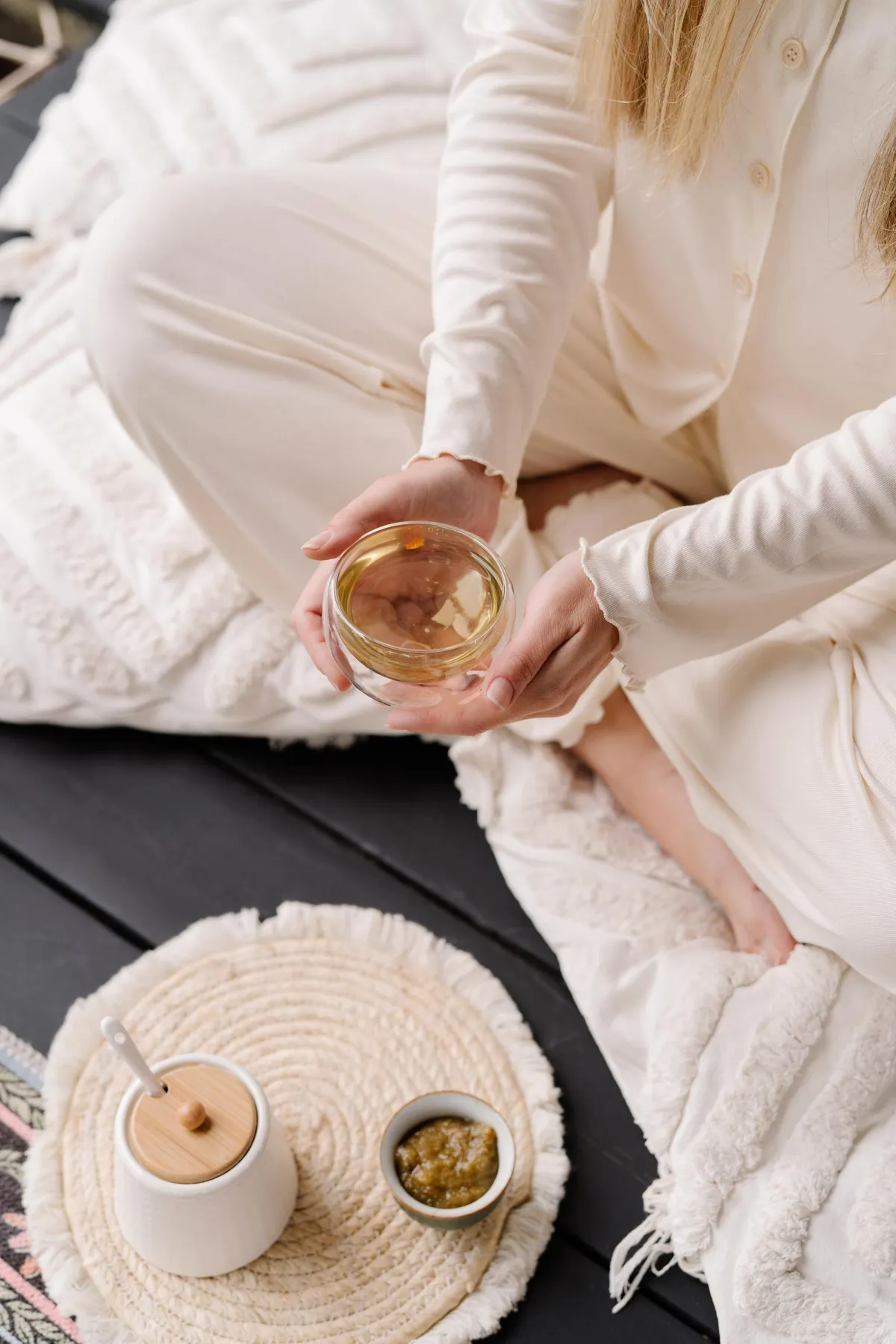 Woman sitting cross-legged on cozy blankets holding warm tea, grounding in a peaceful morning ritual beside natural textures and soft neutral decor
