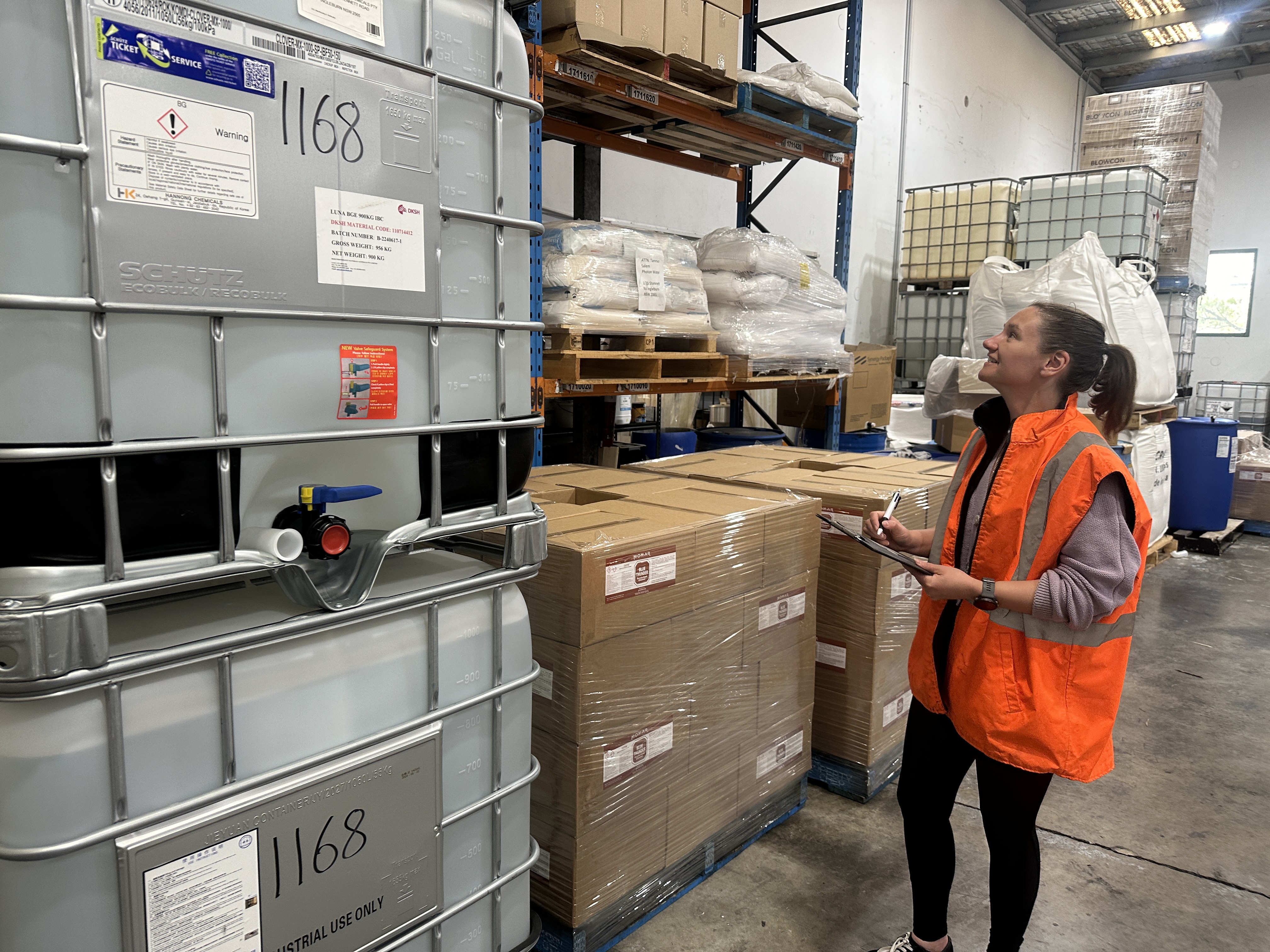 A woman (Sarah Clayton) wearing a bright orange safety vest stands in a warehouse holding a clipboard and pen. She is inspecting large containers and stacks of boxed inventory. The warehouse contains pallets, large tanks, and shelves stocked with various items, suggesting an industrial or manufacturing environment