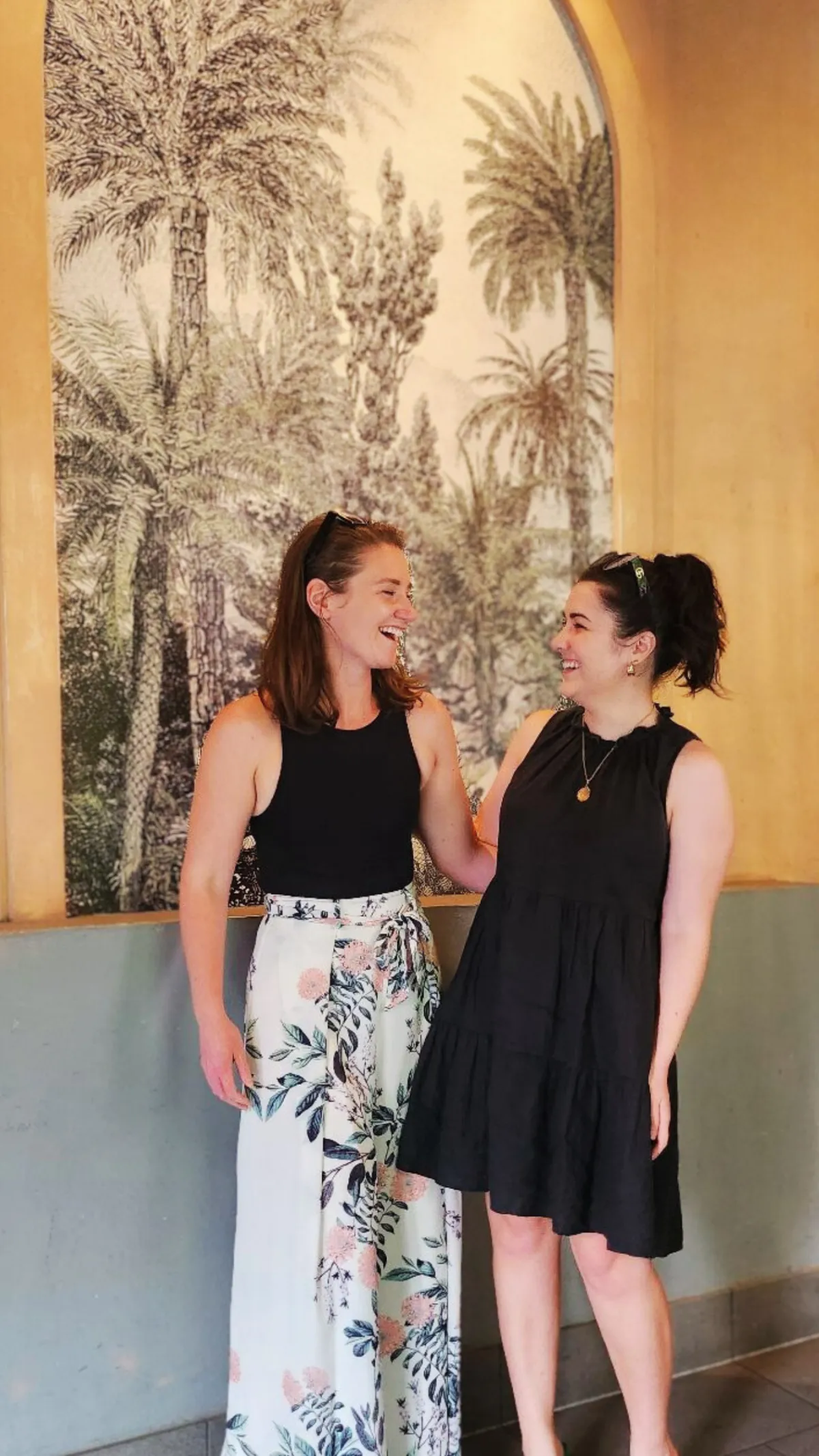 Two women (Sarah Clayton and Aly Nicolaou) standing and smiling at each other in front of a decorative wall featuring a tropical palm tree design. .The setting has warm tones and a relaxed atmosph