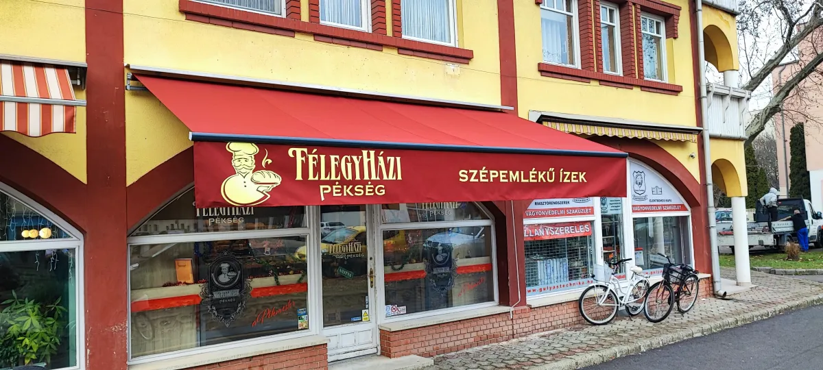 Félegyházi pékség előtt, a járdánál, piros színű napellenző takarja a boltot.