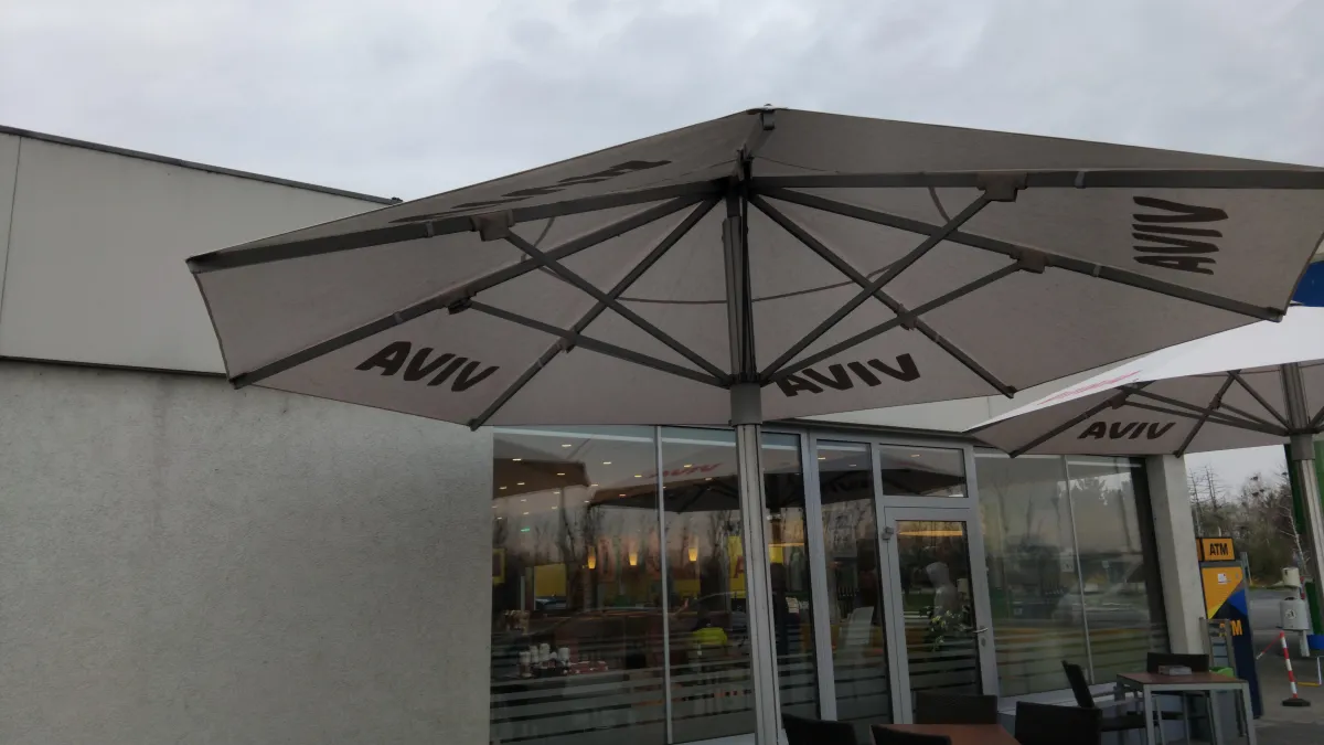 Kávézó terasza szürke napernyőkkel, "AVIV" felirattal, üvegfalú épület előtt, néhány székkel és asztallal, ATM a háttérben, borult ég alatt.