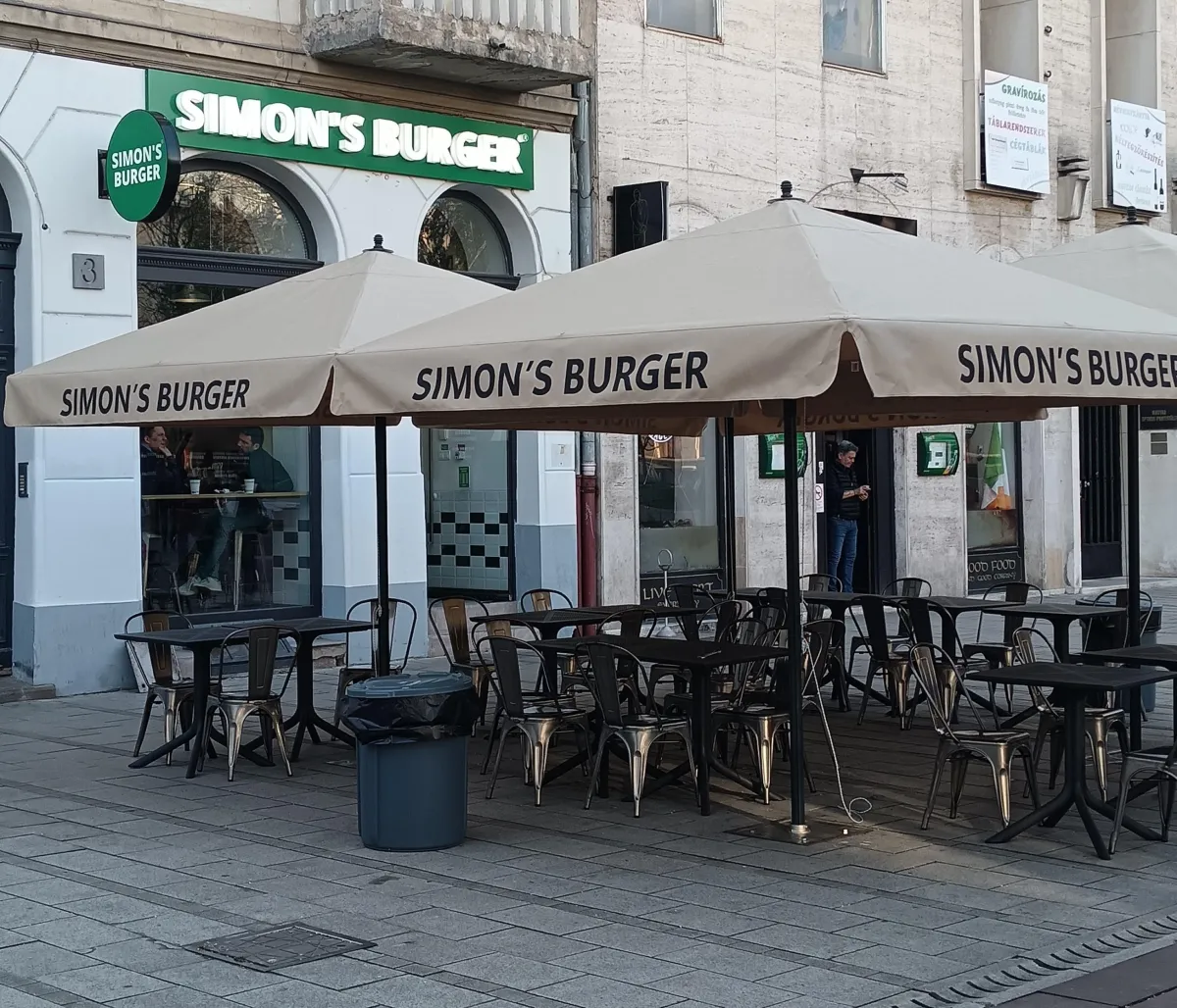 Simon’s Burger étterem terasza bézs napernyőkkel, "SIMON’S BURGER" felirattal, fém székekkel és asztalokkal, néhány vendéggel, kuka a közelben, városi környezetben.