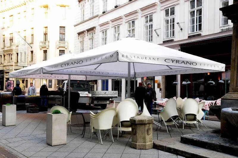 Városi tér egy étterem teraszával, nagy fehér napernyőkkel "CYRANO ÉTTEREM" felirattal, fehér székekkel, emberekkel és klasszikus épületekkel a háttérben.