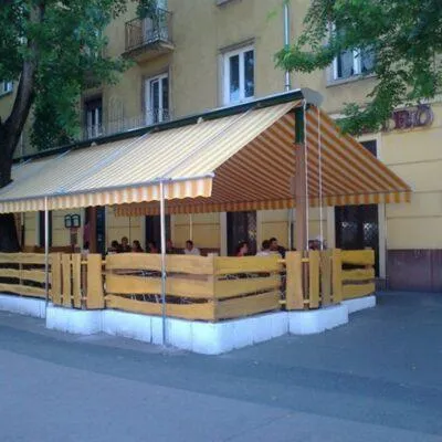 Simon’s Burger étterem terasza bézs napernyőkkel, "SIMON’S BURGER" felirattal, fém székekkel és asztalokkal, néhány vendéggel, kuka a közelben, városi környezetben.