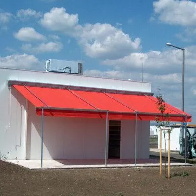 Egy modern fehér épület piros napellenzővel, amely az bejárat fölé nyúlik, árnyékot biztosítva. Az épület lapos tetején egy kis parabolaantenna található. Előtte két kis fa van a földbe ültetve, és egy utcai lámpa áll a közelben. Az ég részben felhős, látható kék foltokkal.