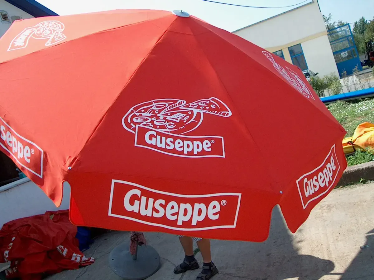 Egy nagy, piros napernyő látható udvari környezetben, rajta többször is a „Guseppe” márkanév fehér betűkkel, valamint pizzaszelet illusztrációk. A napernyő alatt csak két láb látszik, valaki takarásban áll. A háttérben ipari épületek és zöld növényzet figyelhető meg.