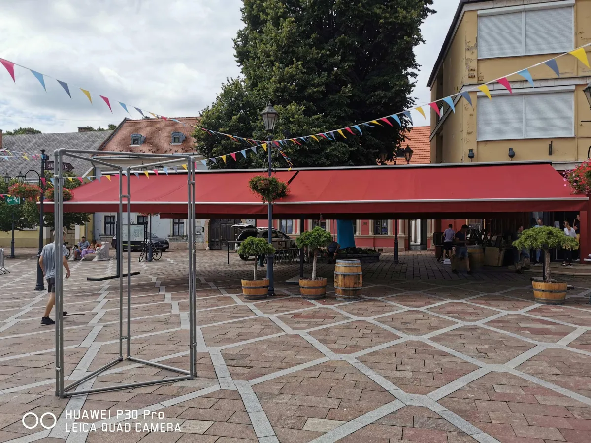 Egy kisvárosi főtéren piros napellenzővel fedett árusítóhely látható, amely alatt néhány ember tartózkodik. A teret színes zászlófüzérek díszítik, körülötte fából készült dézsákban formára nyírt fácskák állnak. Bal oldalon egy fém vázszerkezet, háttérben régi polgárházak, jobbra sárga vakolatú, redőnnyel zárt ablakos épület látható. A tér burkolata világos mintás kő. Az egész helyszín egy hagyományos, rendezett főtéri piaci vagy közösségi eseményre utal, nyugodt, nyári délutáni hangulatban.