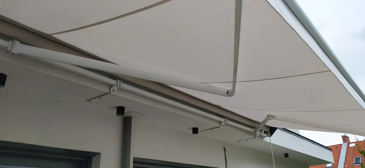 Egy modern, fehér napellenző látható egy családi ház teraszán. A napellenző fémből készült váza az épület homlokzatához van rögzítve, a szerkezeti karok nyitott állapotban tartják a kifeszített ponyvát. A vászon enyhén ívelt, világos színű, vékony barna csíkok díszítik. A mennyezetről leeresztett napellenző kellemes árnyékot biztosít a terasz alatt, a háttérben részben látható a szomszédos ház piros cserepes teteje és az ég is. Az egész jelenet napos, rendezett, modern környezetet sugall.