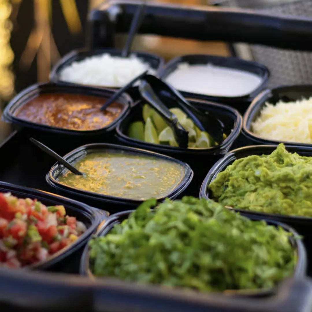 A variety of Mexican food toppings including salsa, guacamole, shredded lettuce, and limes in black containers.