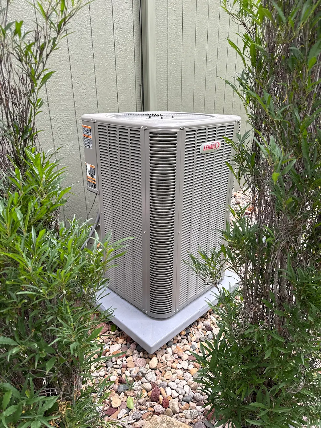 Exterior shot of a newly installed Lennox central air conditioning condenser unit sitting on a fresh plastic pad and clean gravel base next to vinyl siding.