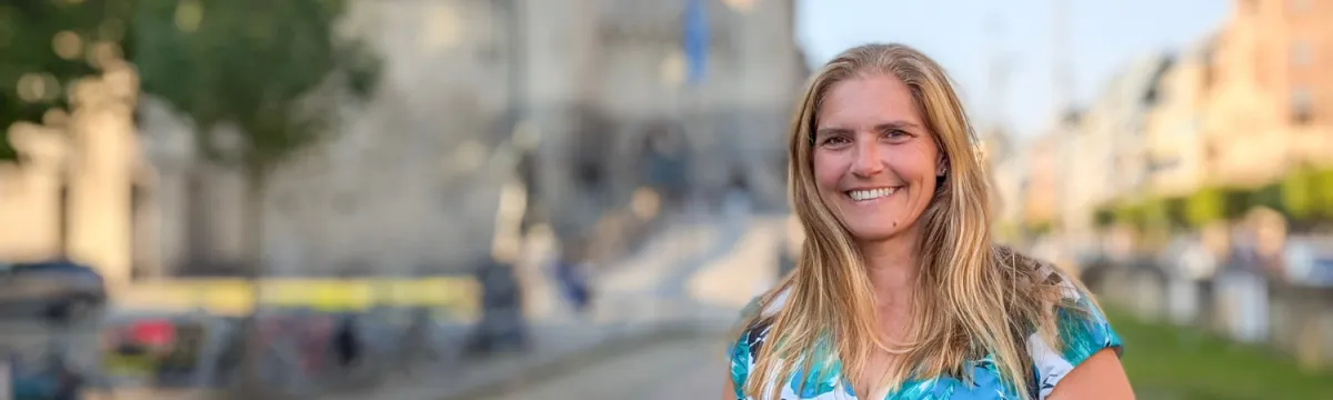 Jennifer Guerrero smiling outdoors in a blue patterned dress, with a softly blurred streetscape and stone building behind her.