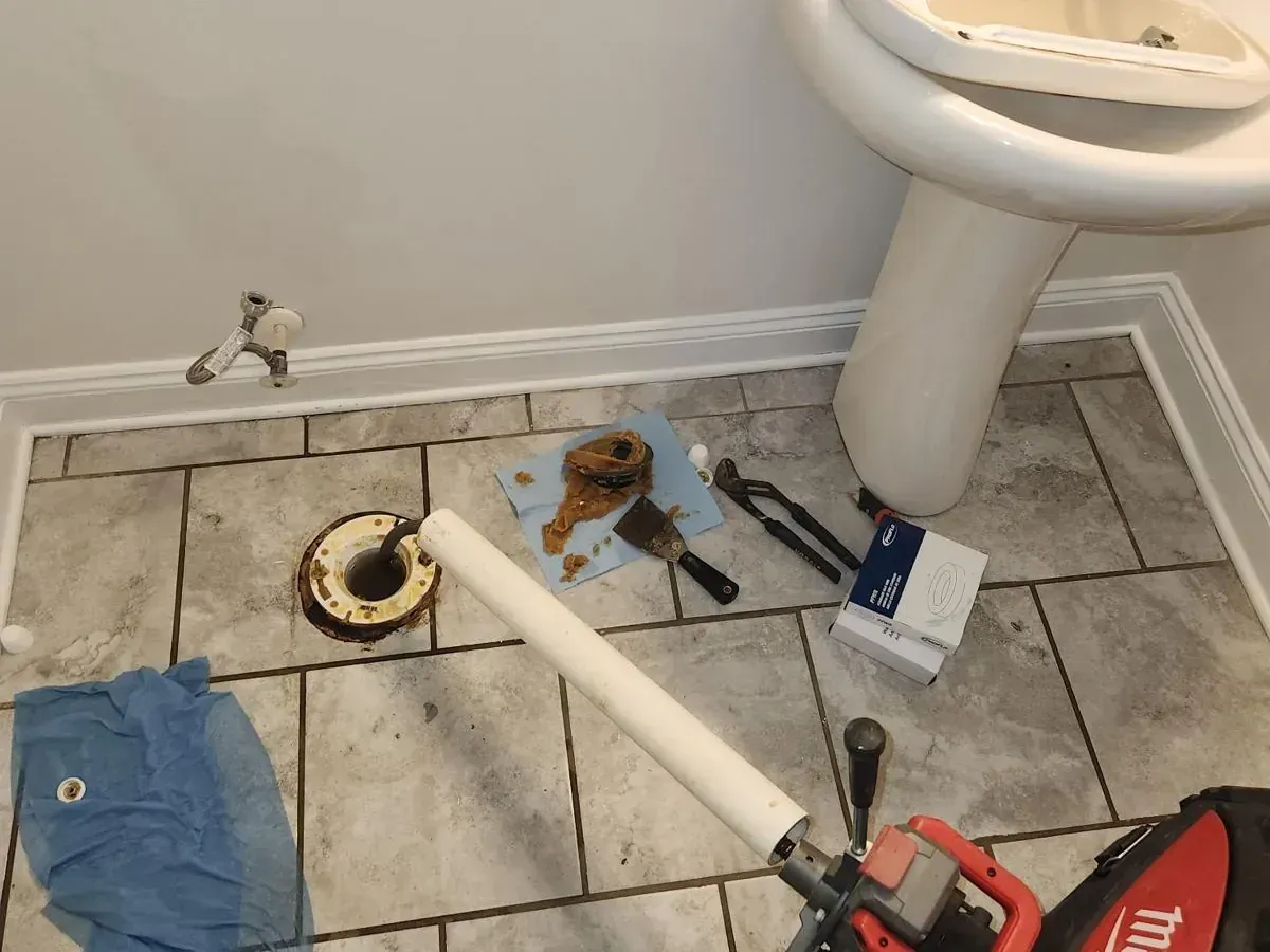 Bathroom toilet flange repair in progress with plumbing tools, removed toilet, drain auger, and exposed flange on tile floor.