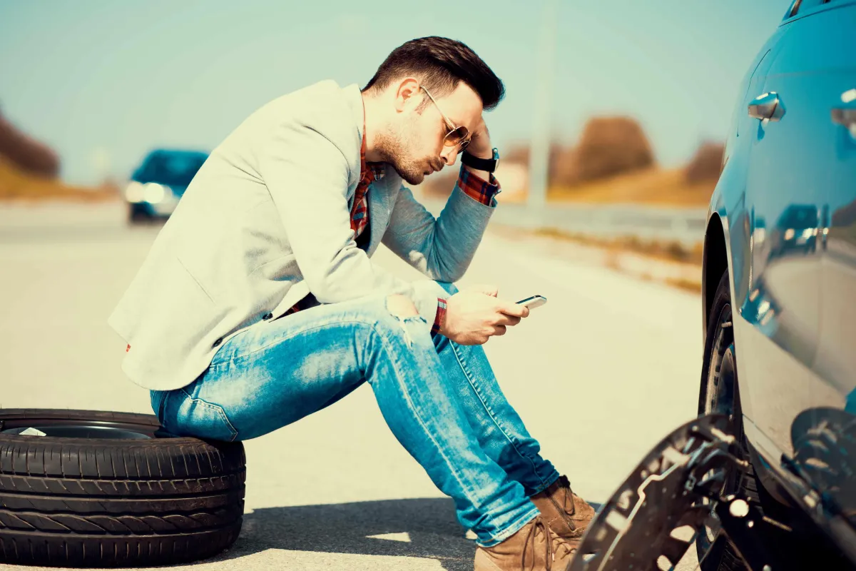 Man sitting on a tire next to a broken car looking at a phone screen