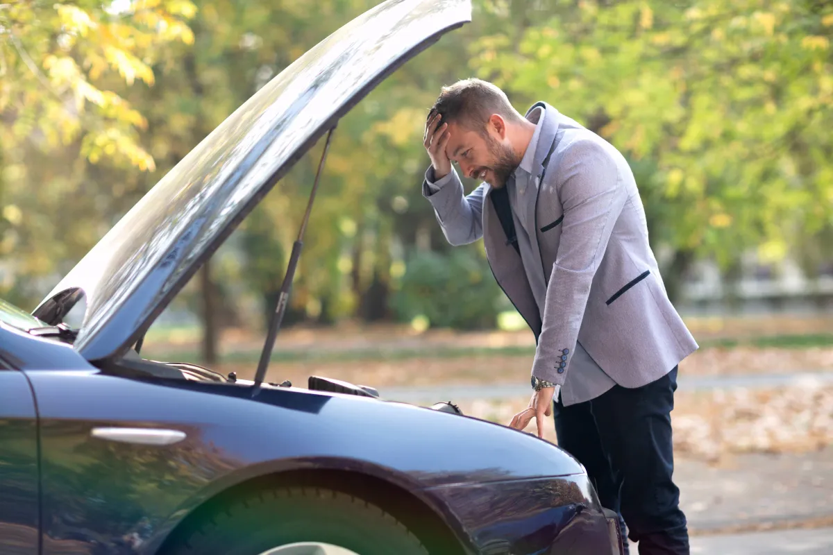 Man with hand on his forehead, looking inside the hood of a broken down car