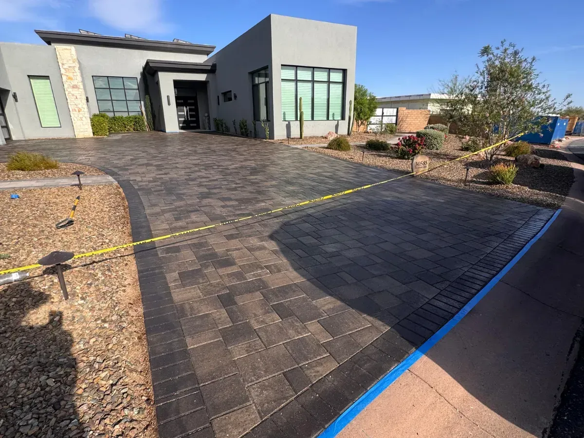 A modern gray house with a new dark gray paver driveway, cordoned off by yellow caution tape under a clear sky.