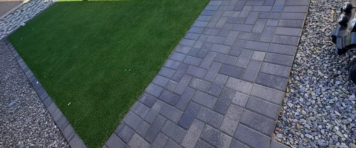 A diagonal view of artificial green grass, dark gray herringbone paver walkway, and a gray gravel area.