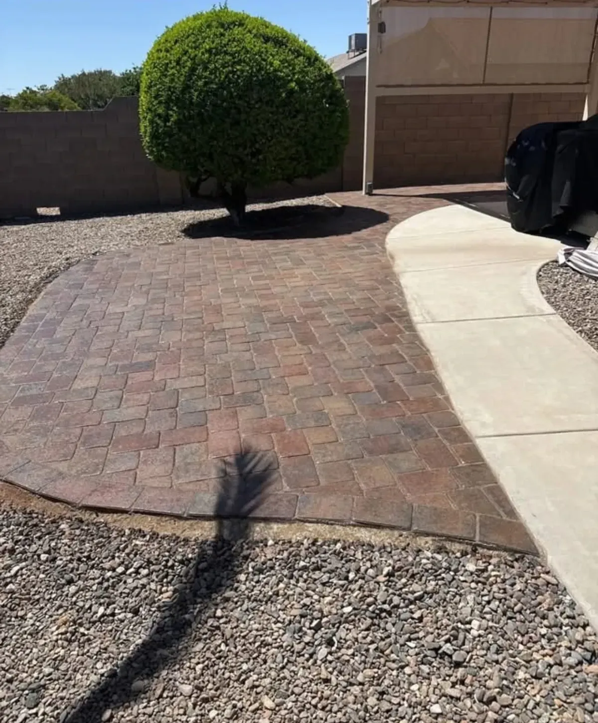 A backyard with a textured paver patio, a manicured green bush, gravel, and a curved concrete walkway on a sunny day.