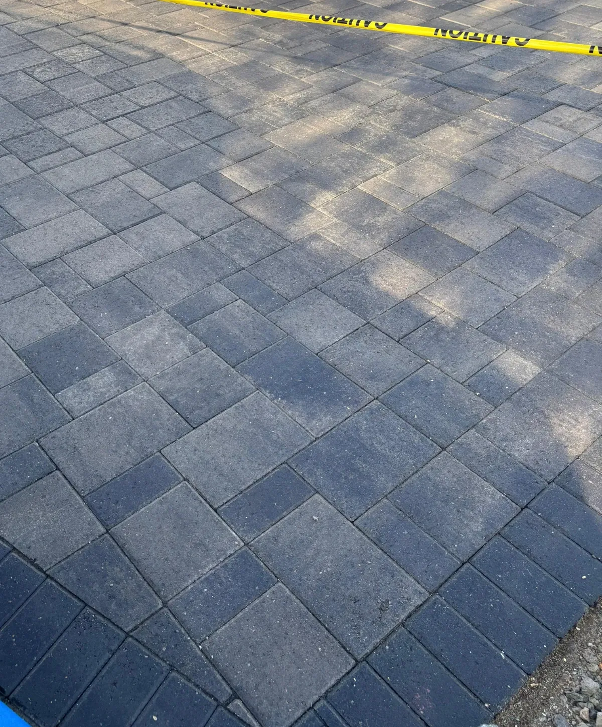 Dark gray interlocking pavers forming a patterned surface with yellow caution tape stretched across the top.