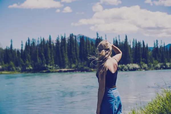 A woman with long, dark hair gazes out over a serene, blue river bordered by a forest under a partly cloudy sky.