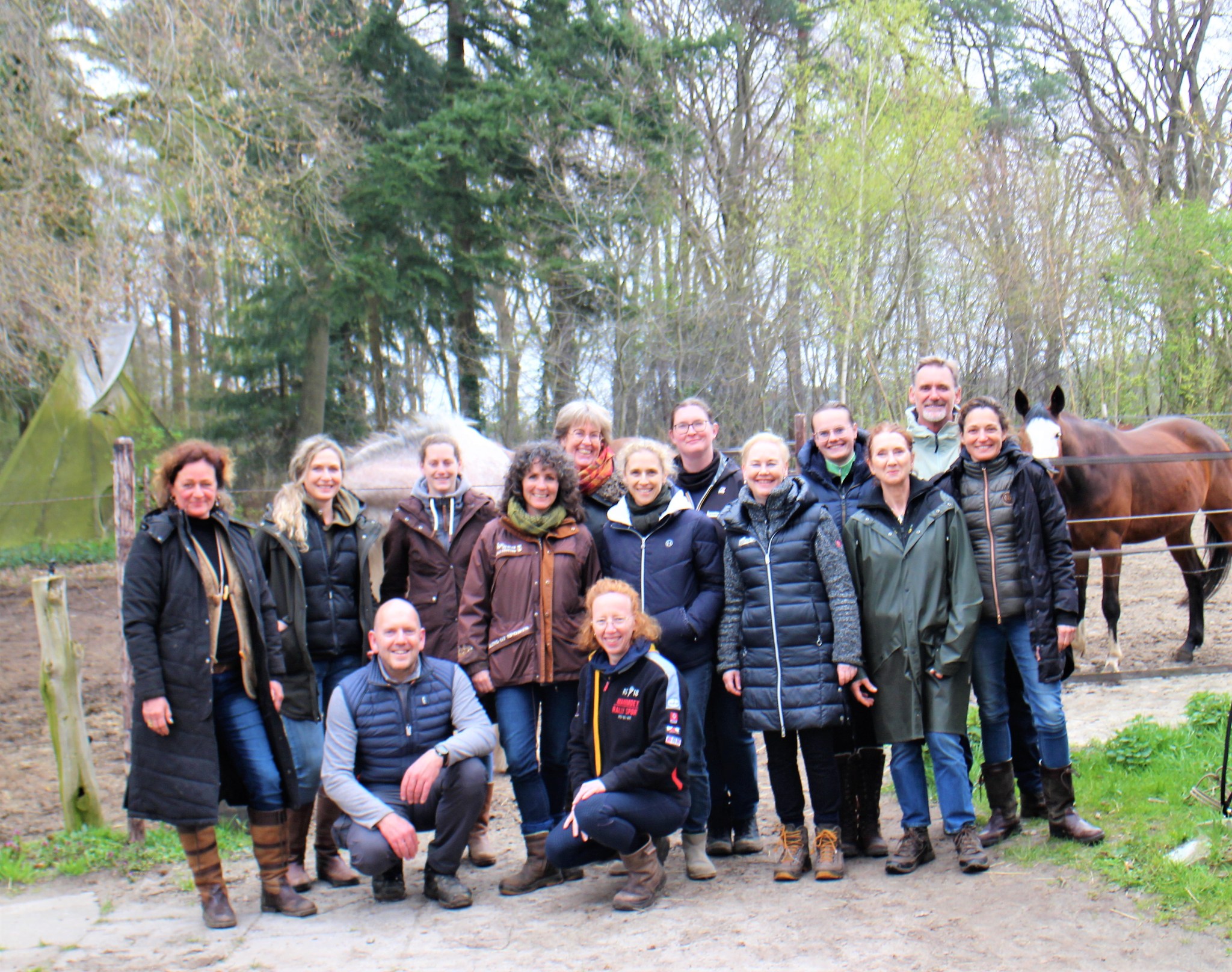 A diverse group of equestrians, men and women of various ages, standing in a circle with their horses in a golden field, hands joined, smiling and radiating unity and accomplishment. The scene is bathed in warm, late afternoon light.