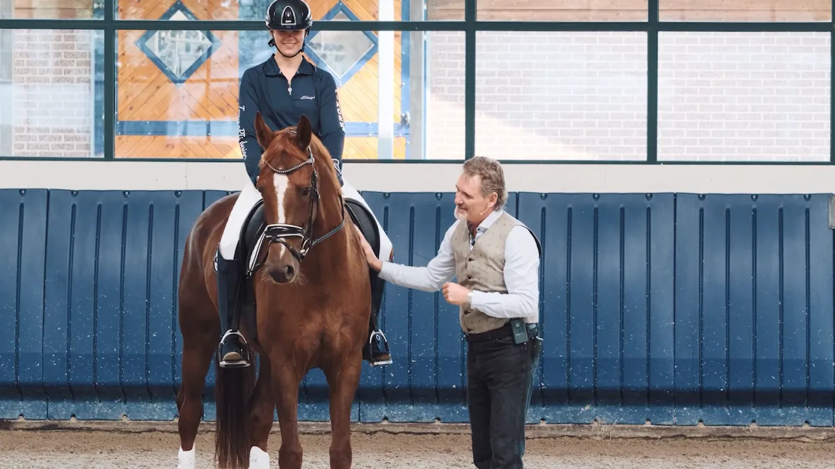 A focused female equestrian in mid-ride, guiding a bay horse through a precise dressage movement in a sunlit, airy indoor arena. The rider wears a tailored jacket and helmet, exuding confidence and technical skill. The background is softly blurred, emphasizing the harmony between horse and rider.