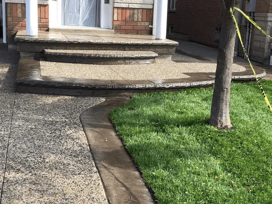 Front porch and walkway made of exposed aggregate concrete with a smooth, dark-stamped border. The curved pathway blends neatly into the grass lawn, adding polished charm to the home's exterior.