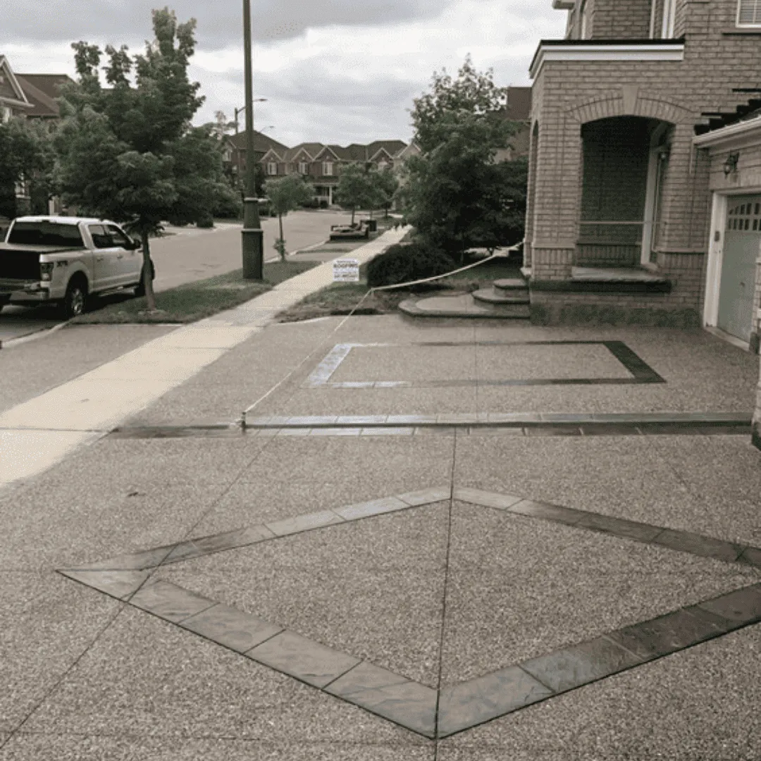 Exposed aggregate concrete driveway with dark decorative border inlays forming square and diamond patterns. A clean and structured look that highlights custom concrete work for residential driveways.