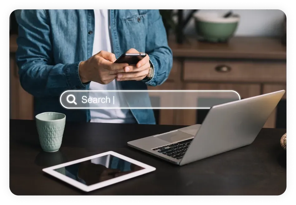 Person using a smartphone with a laptop and tablet on the table, accompanied by a virtual search bar overlay, highlighting the role of SEO services in Dallas for optimizing online visibility and search rankings.