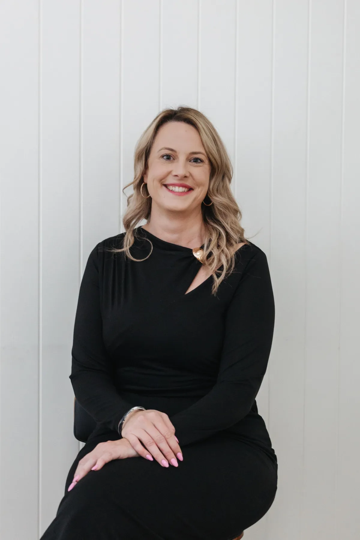 Cristina, a medical receptionist at Labiaplasty Sydney, smiling in a black dress, seated against a white wooden backdrop, embodying warmth and professionalism in women's health care.